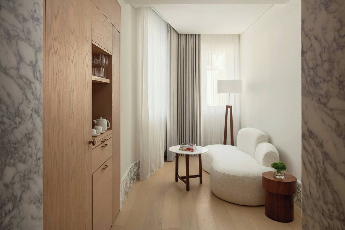A compact hotel room with a wooden wardrobe, a round table, a white armchair, a small side table with a plant, and a floor lamp.