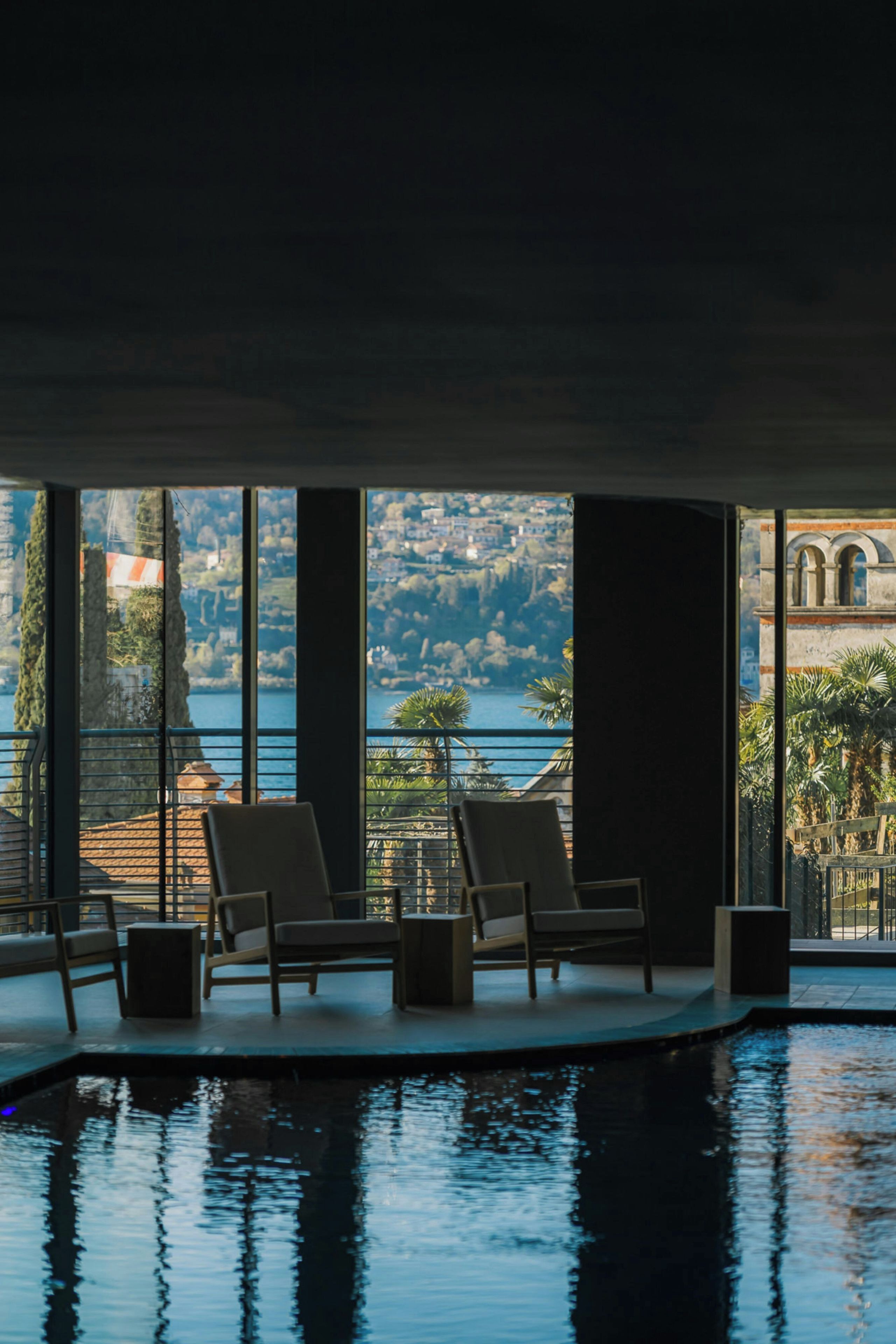 Ein Poolbereich mit zwei Stühlen und einem kleinen Tisch, große Fenster mit Blick auf einen See und Gebäude.