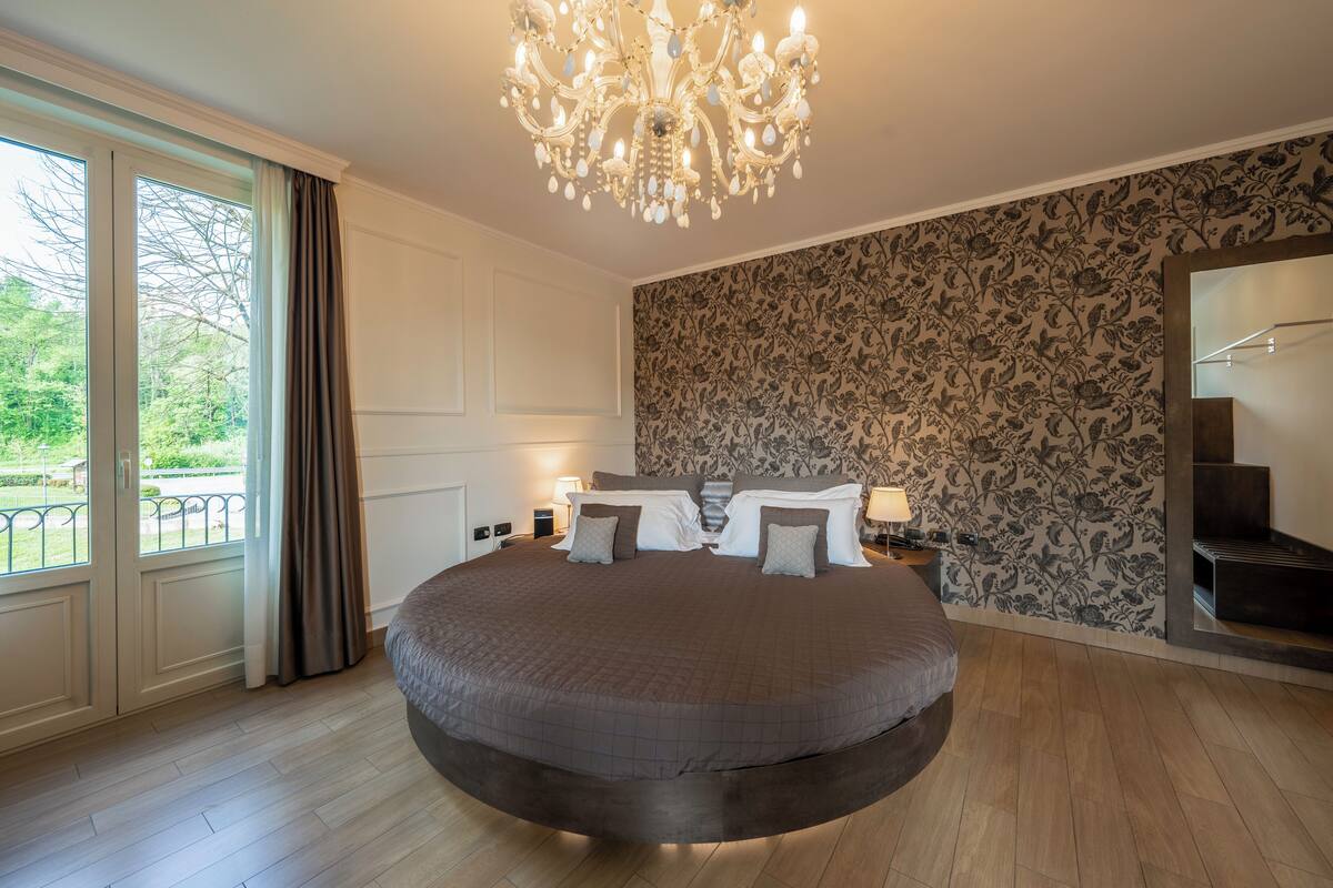 A bedroom with a round bed, a chandelier, a view of a garden, and a patterned wallpaper.