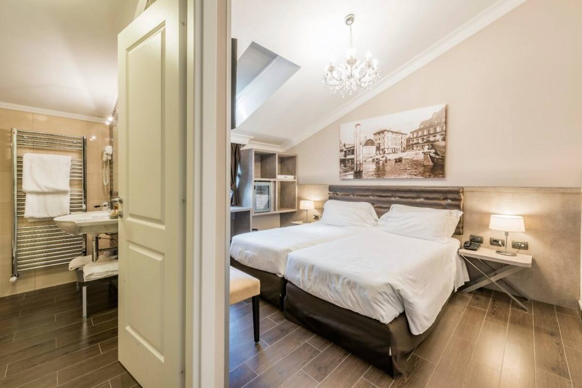 A hotel room with a large bed, a bedside table with a lamp, a bathroom with a towel rack, and a cityscape painting on the wall.