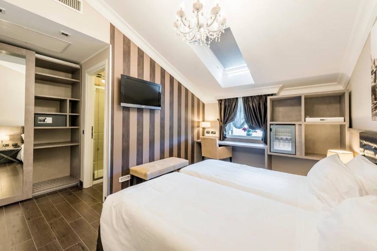 A hotel room with a bed, a TV, a desk, a chair, a bench, and a wardrobe.