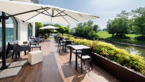 Een terras buiten met tafels en stoelen, een overdekt gedeelte met parasols en uitzicht op een rivier en groen.