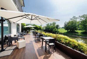 An outdoor patio with tables and chairs, a covered area with umbrellas, and a view of a river and greenery.