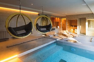 A modern indoor pool area with two hanging chairs, lounge chairs, and a hot tub.