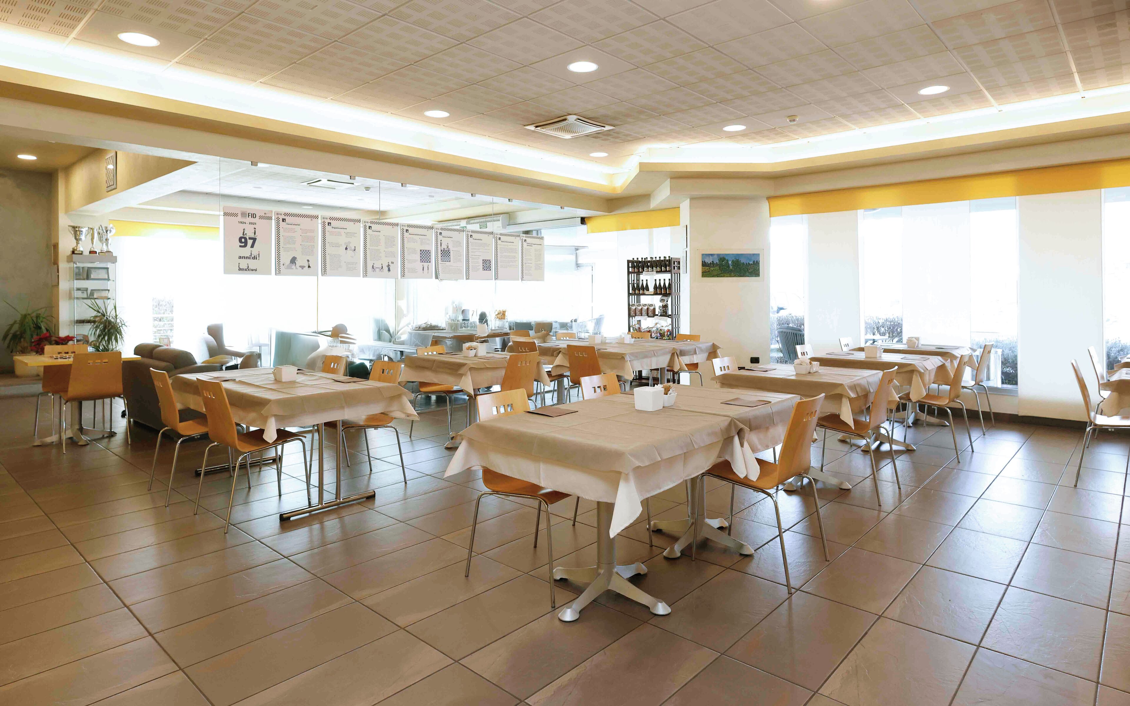 A spacious dining area with multiple tables and chairs, a ceiling with recessed lighting, and a view of the outdoors through large windows.
