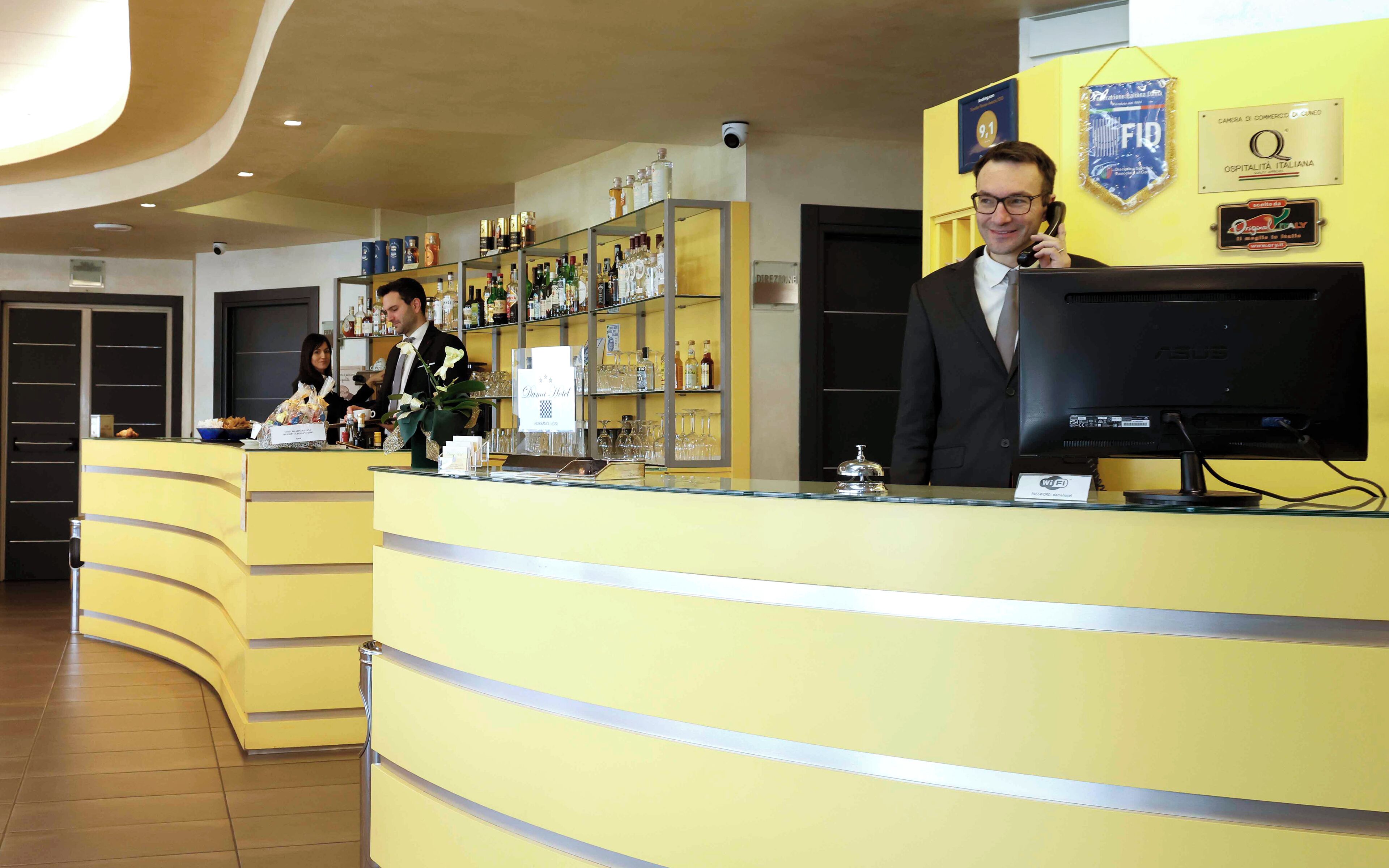 Zona de recepción de hotel con un mostrador amarillo, un monitor de computadora y diversos servicios, incluyendo un minibar y una vitrina de vidrio.