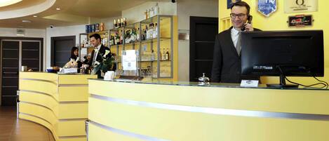 A hotel reception area with a yellow counter, a computer monitor, and various amenities including a minibar and a glass display.