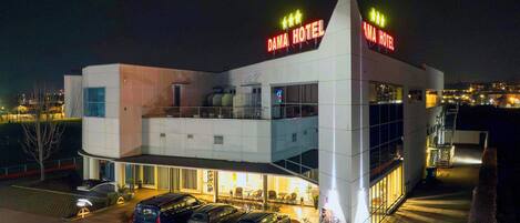 Un hotel moderno con un llamativo letrero que dice 'DAMA HOTEL', un estacionamiento con varios coches y una entrada bien iluminada.