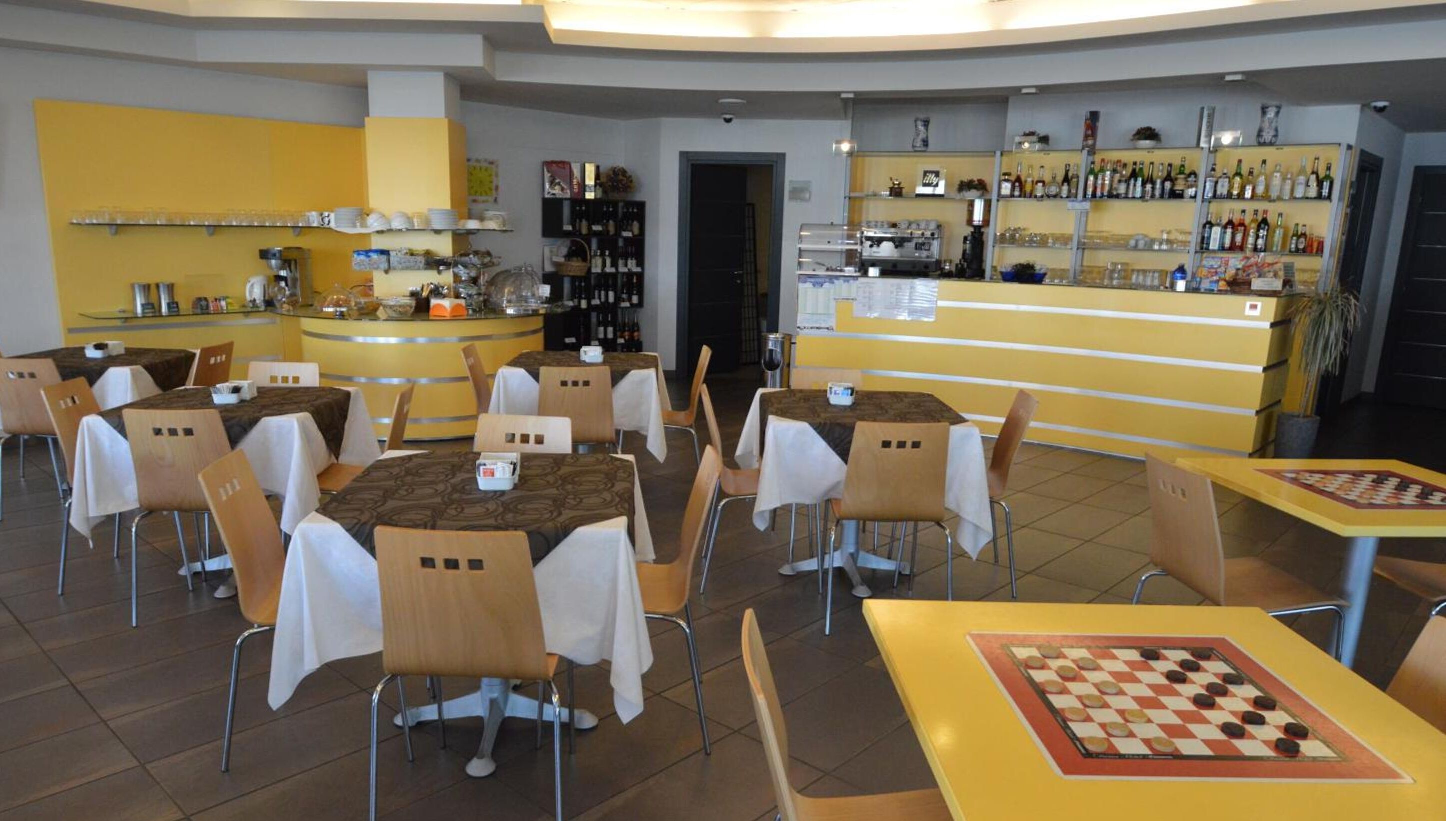 A restaurant with multiple tables and chairs, a bar area with shelves of bottles, and a chess table.
