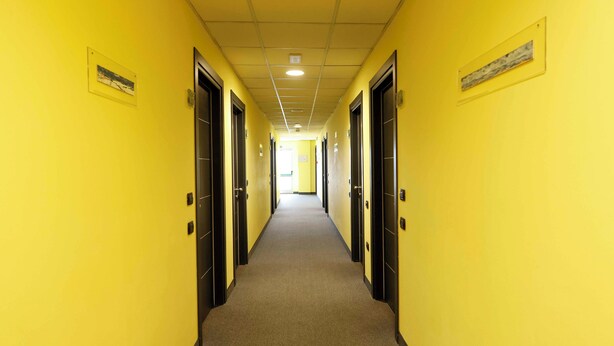 A long, narrow hallway with yellow walls and black doors, leading to a bright room at the end. - Hotel Dama (Fossano)