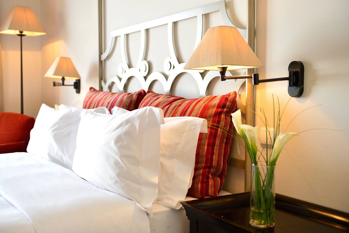 A neatly made bed with white and red striped pillows, a bedside table with a vase of flowers, and wall-mounted lamps.