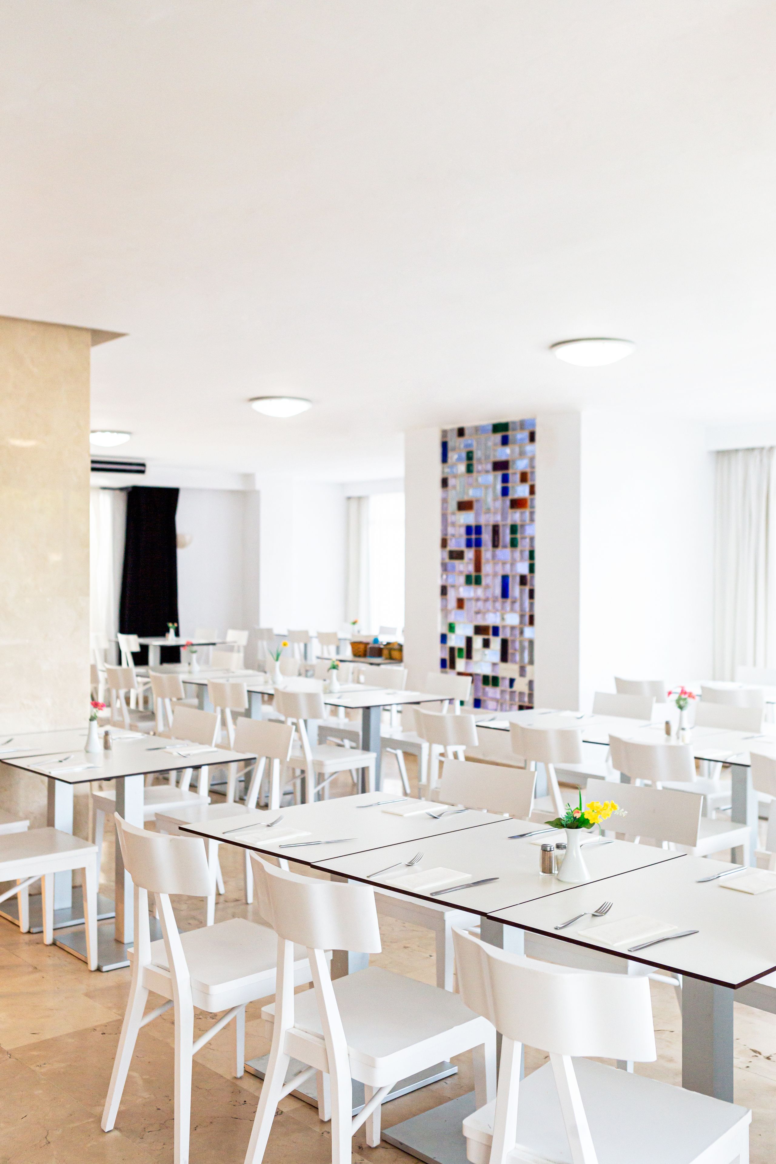 A modern dining area with white tables and chairs, a colorful mosaic wall, and a clean, minimalist design.