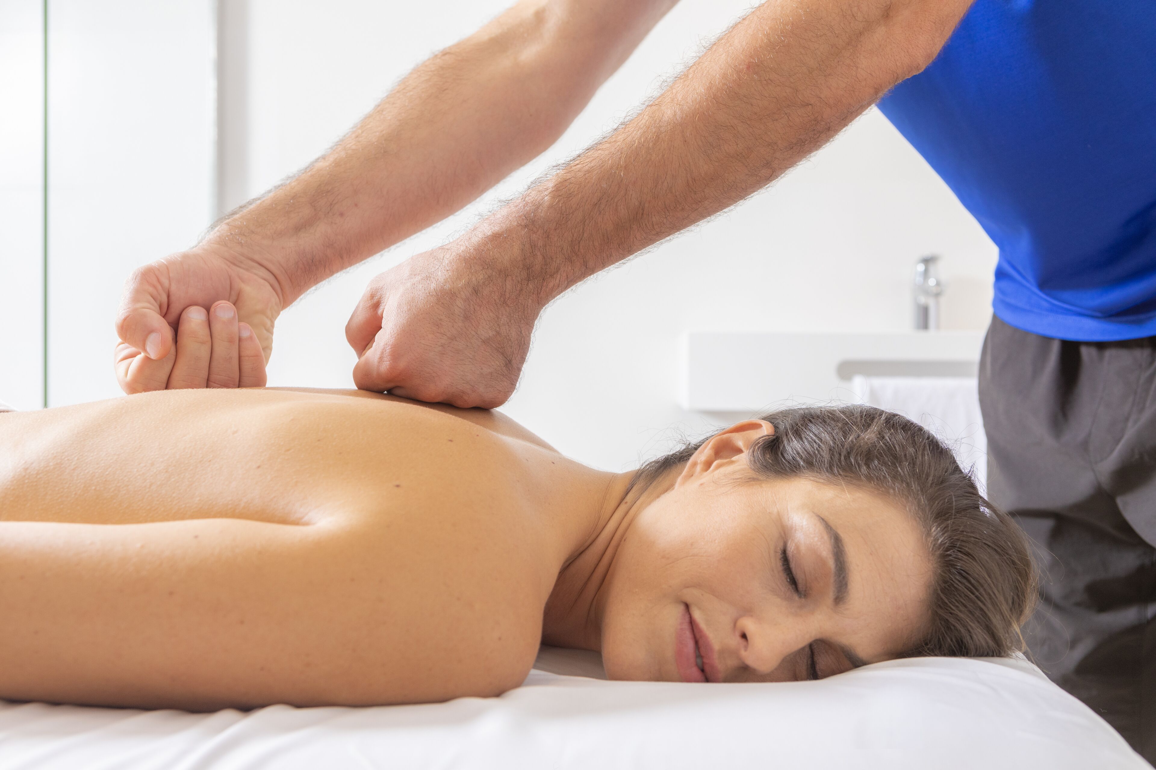 A person receiving a massage on a bed in a room with a white wall and a small window.