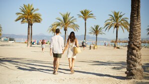 A couple walking on a sandy beach with palm trees and mountains in the background. - Hotel JS Paradise Sport - Adults Only (Llucmajor)