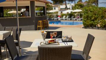 A dining area with wicker chairs and tables, a pool in the background, and a bar area with umbrellas.