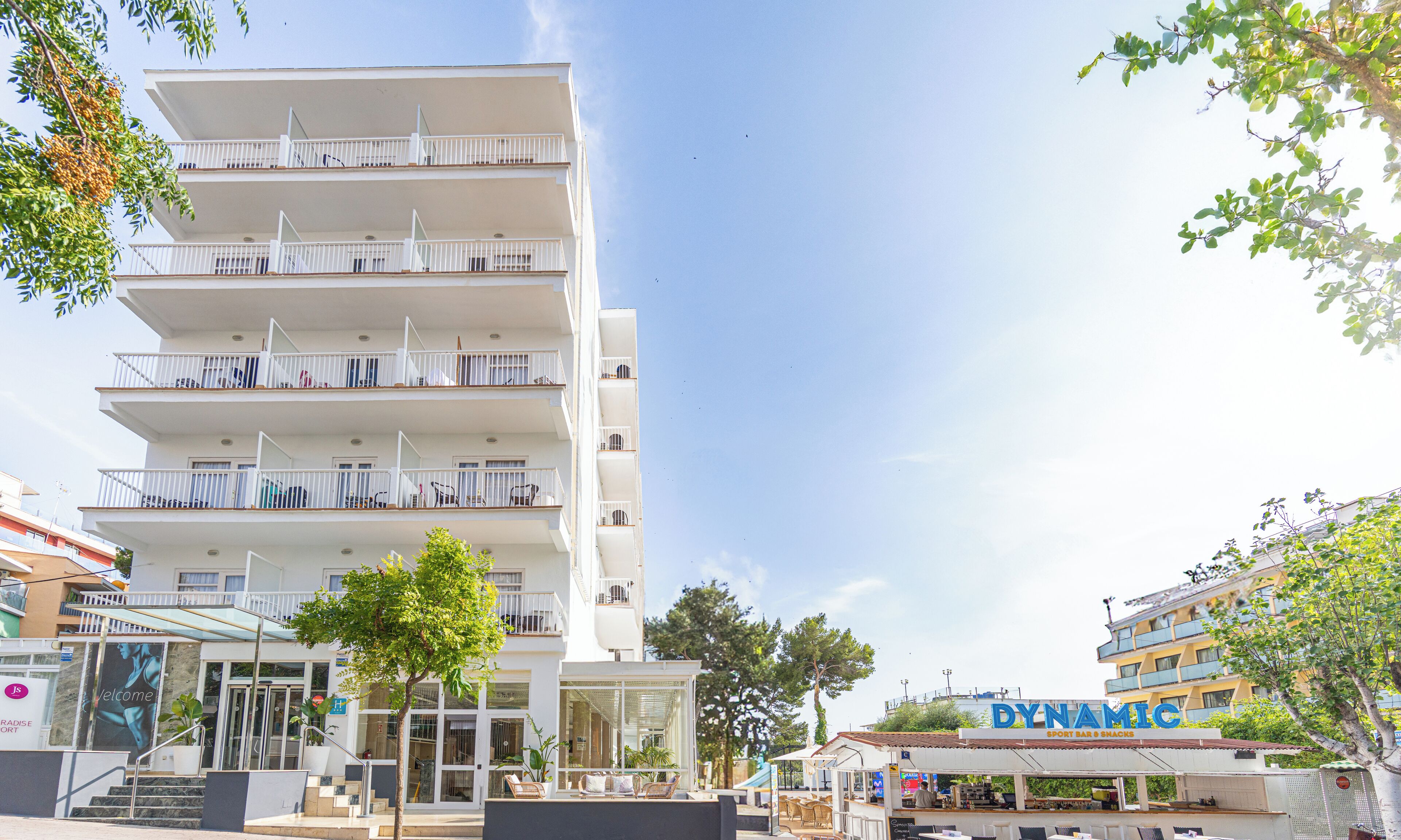 Ein mehrstöckiges Hotel mit Balkonen, ein Restaurant namens 'DYNAMIC' im Vordergrund und ein 'Welcome'-Schild am Eingang.