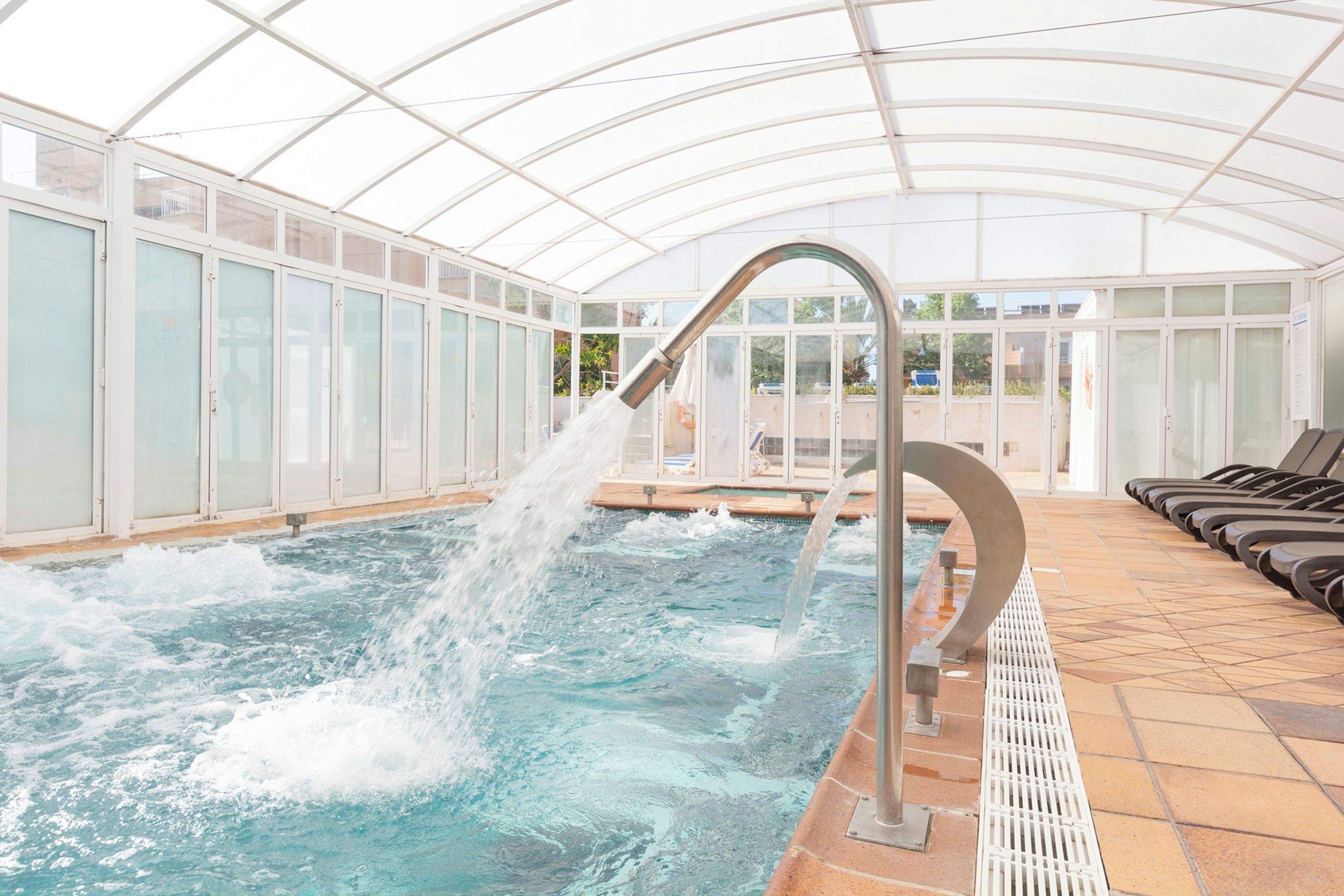 A swimming pool with a water feature, surrounded by glass walls and a tiled floor.