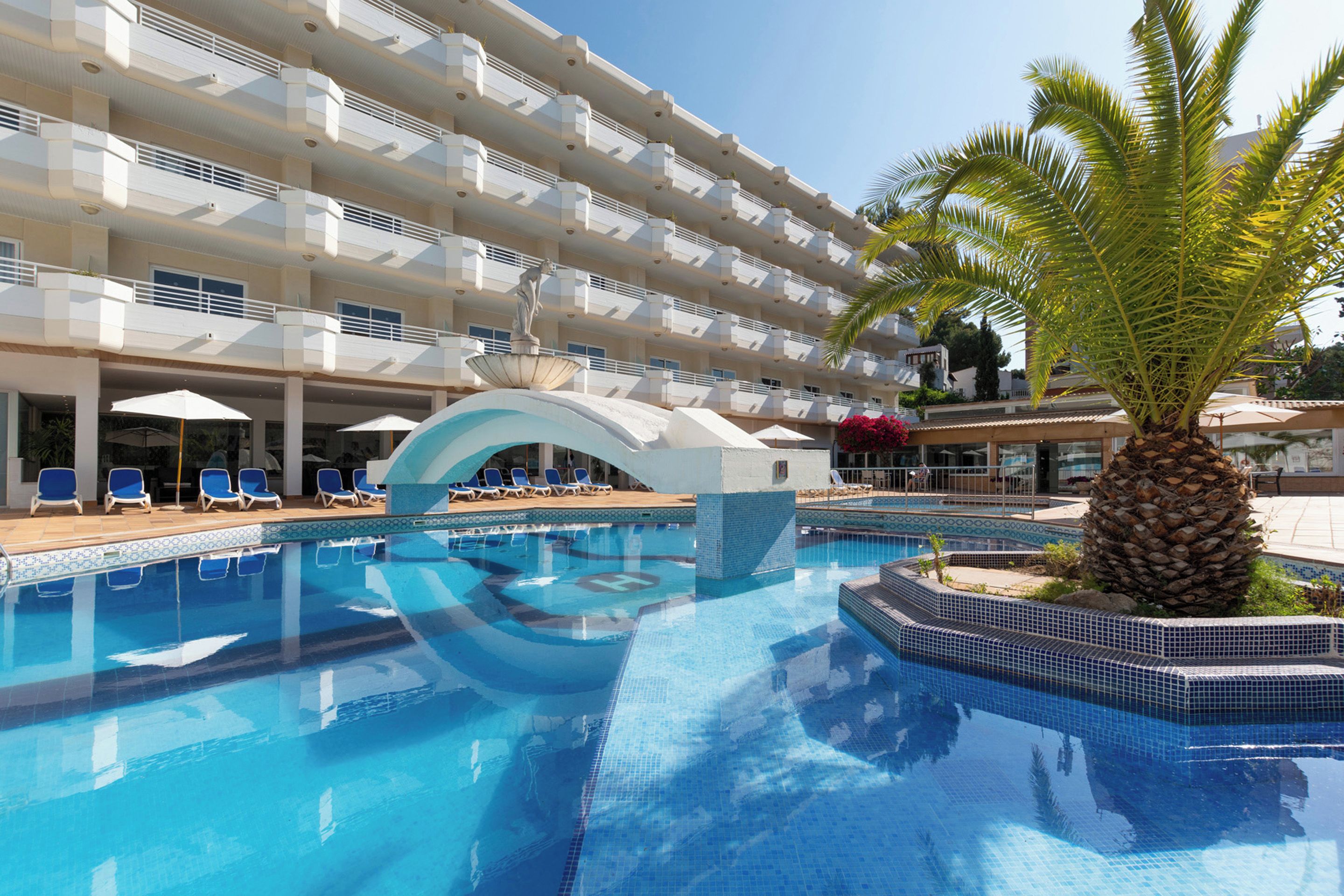 A hotel pool area with multiple levels of balconies, a central fountain, and a palm tree.