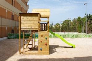 Una estructura de juego de madera con agarres para escalar y un tobogán, ubicada sobre suelo arenoso con una cerca verde y edificios al fondo.