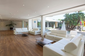 A spacious hotel lobby with a modern design, featuring a large seating area with white sofas and a wooden coffee table.