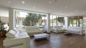 Een ruime woonkamer met meerdere witte banken en een salontafel, grote ramen met uitzicht op palmbomen en geparkeerde auto's.