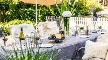 Een terras met een eettafel gedekt voor twee personen, met een wijnfles, glazen, een kaasplankje en een vaas met bloemen.