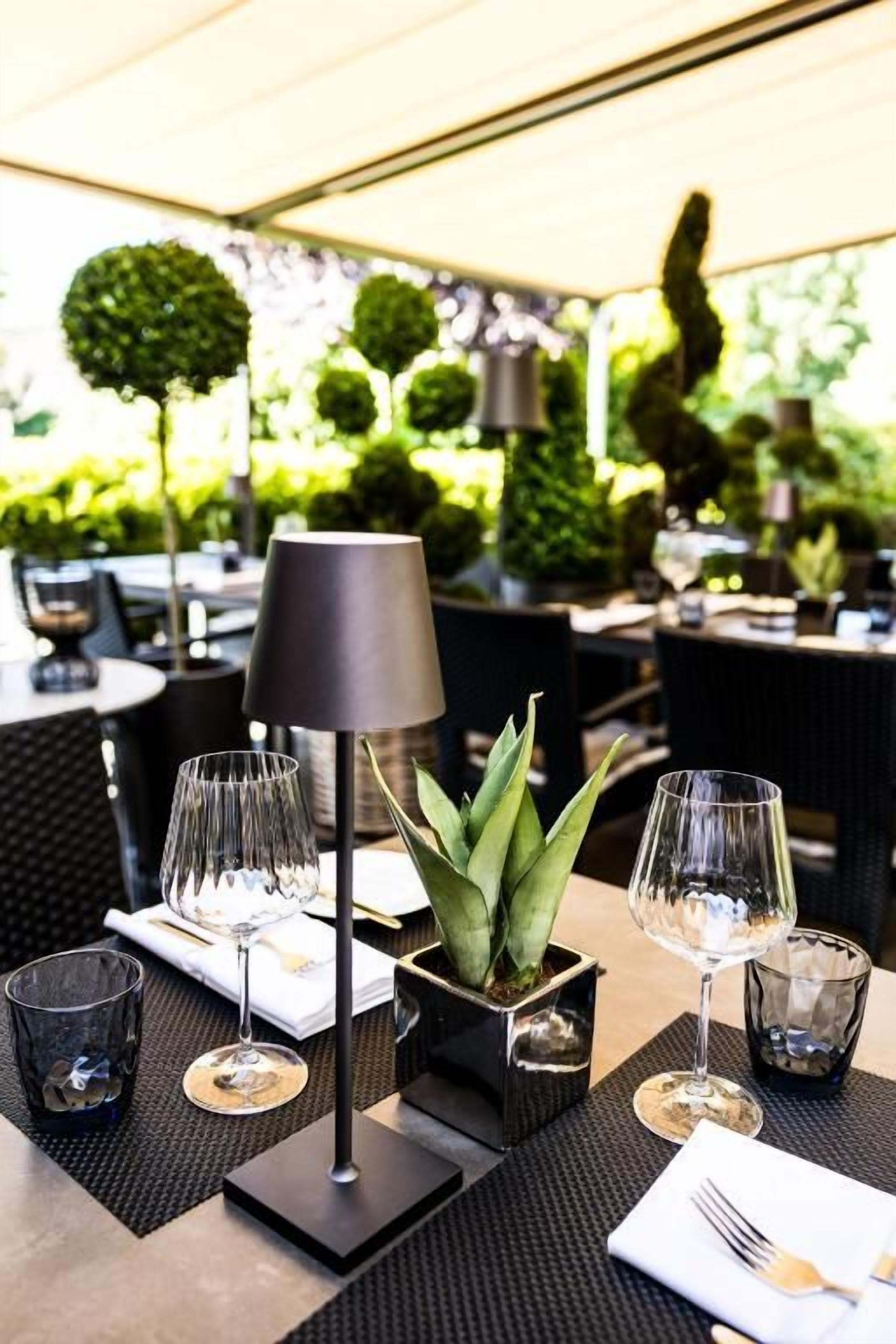 Een buiten eethoek met tafels voor twee personen, met zwarte tafelkleden, transparant servies en een centrale potplant.