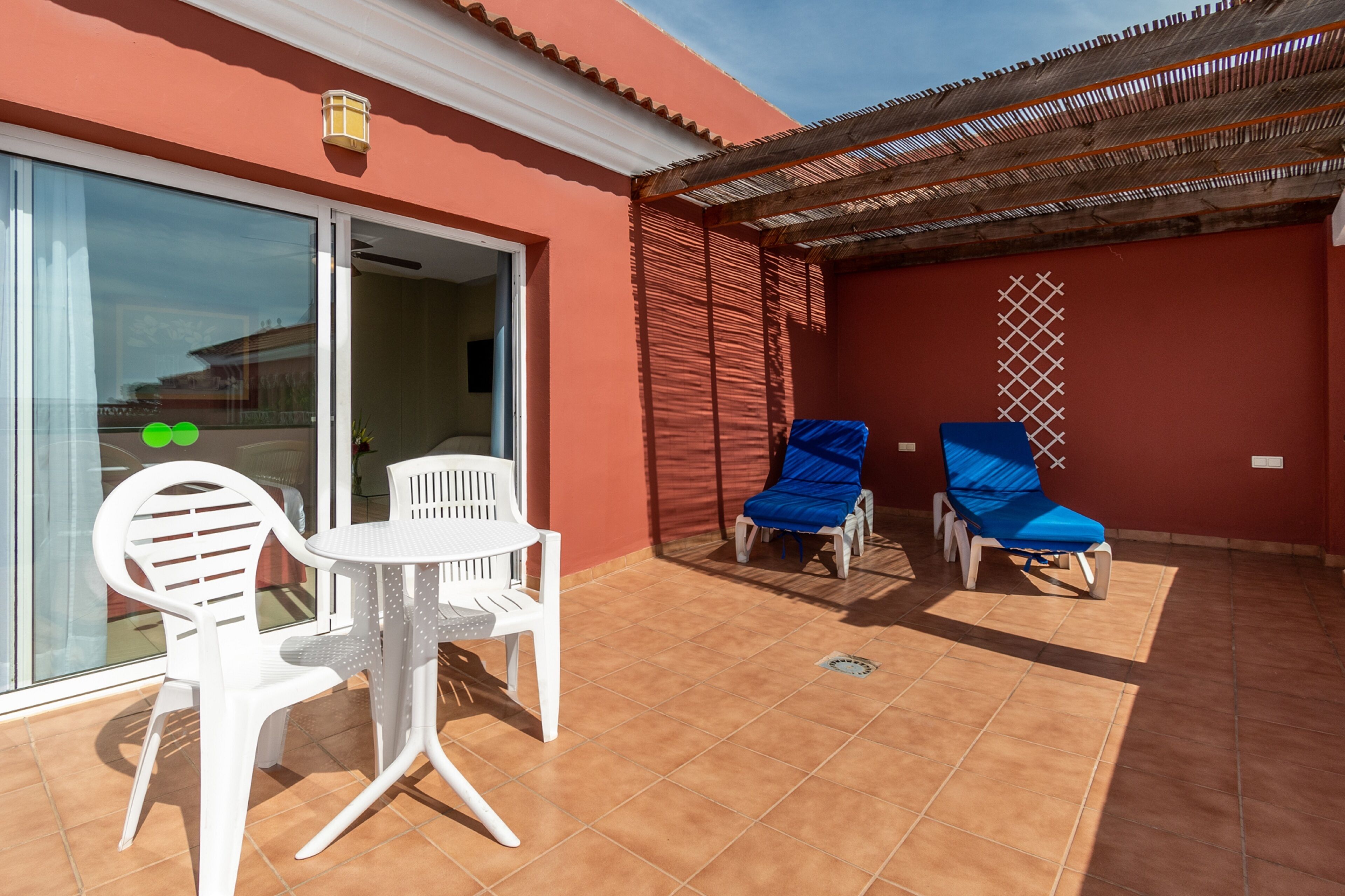 Un patio con sillas de plástico blanco y una mesa, una zona techada con sillas azules y una pared roja con una ventana.