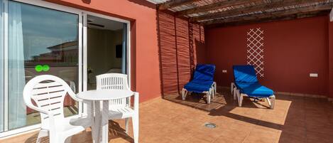 Un patio con sillas de plástico blanco y una mesa, una zona techada con sillas azules y una pared roja con una ventana.
