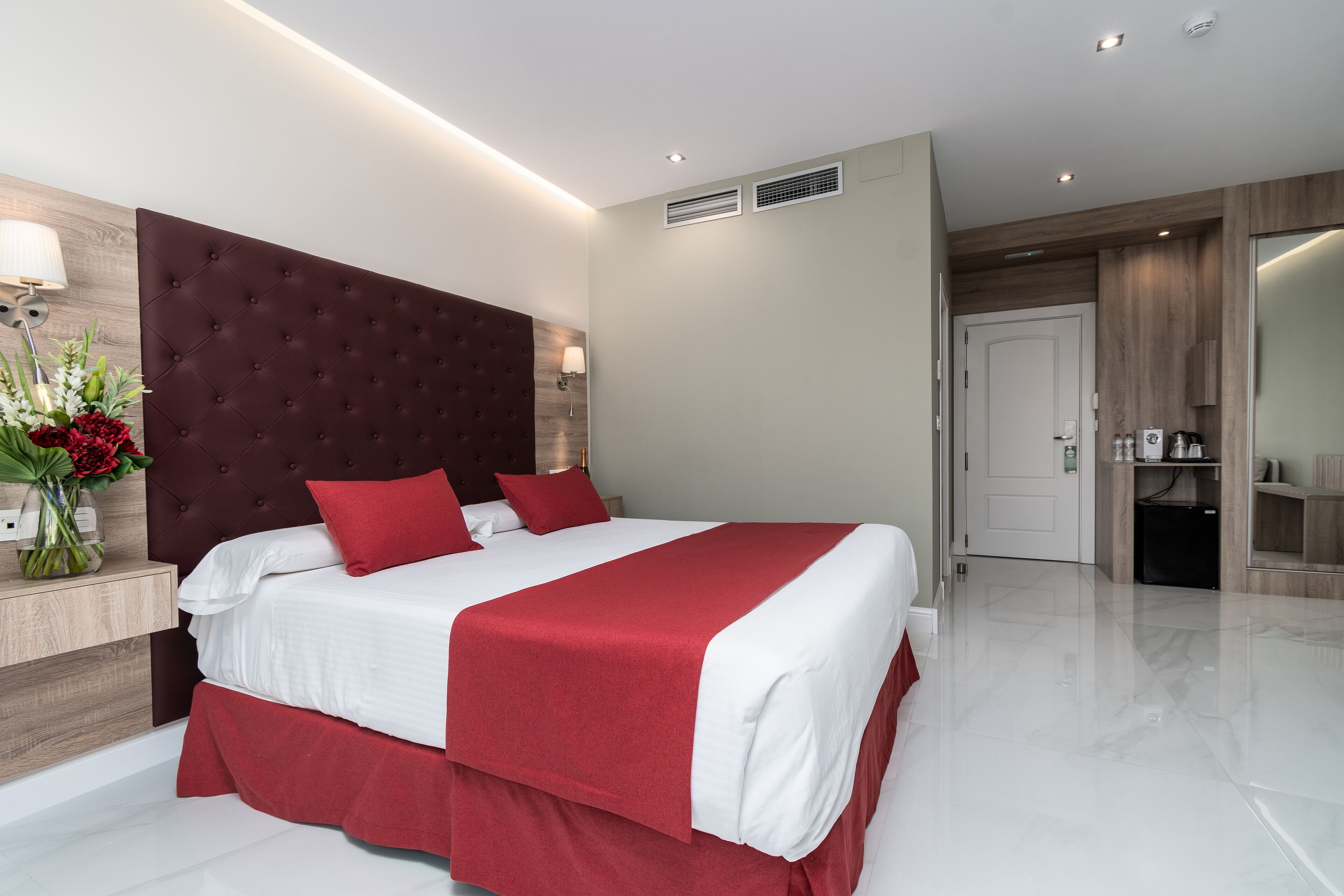 A modern hotel room with a large bed, a wooden headboard, a bedside table with a lamp, and a small kitchenette.