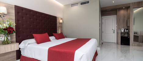 A modern hotel room with a large bed, a wooden headboard, a bedside table with a lamp, and a small kitchenette.