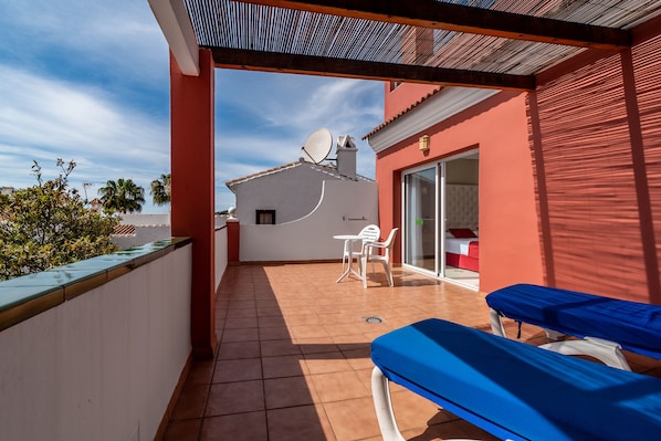 Un balcón con dos sillones azules, una mesa blanca y un ventanal de vidrio que da a una habitación interior.