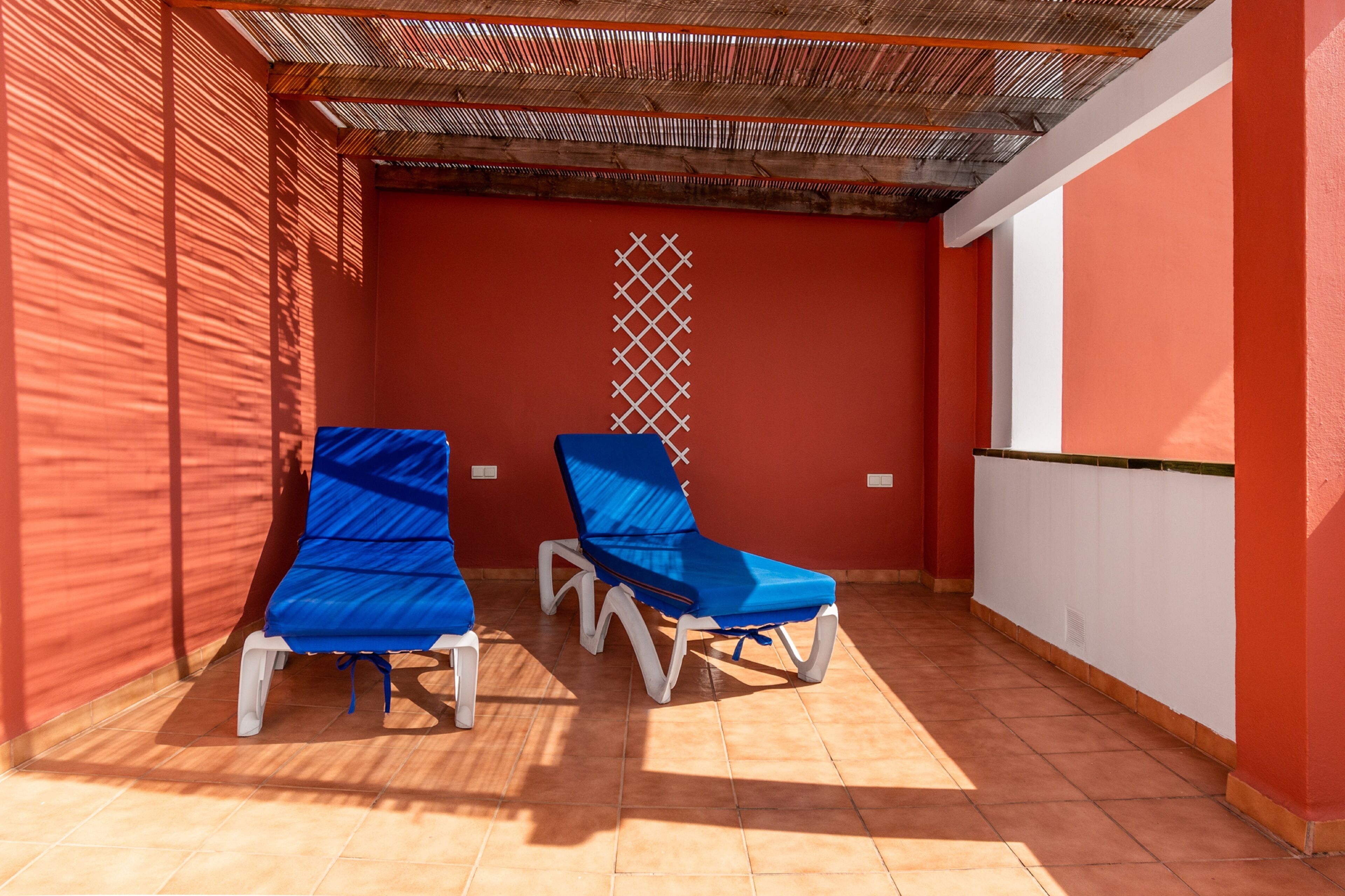 Una habitación iluminada por el sol con dos sillones azules, una pared roja y una partición blanca con diseño de celosía.