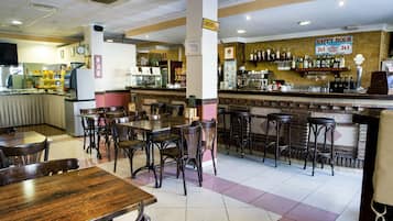 Un interior de restaurante con mesas y sillas de madera, una zona de bar con taburetes y un reloj en la pared.
