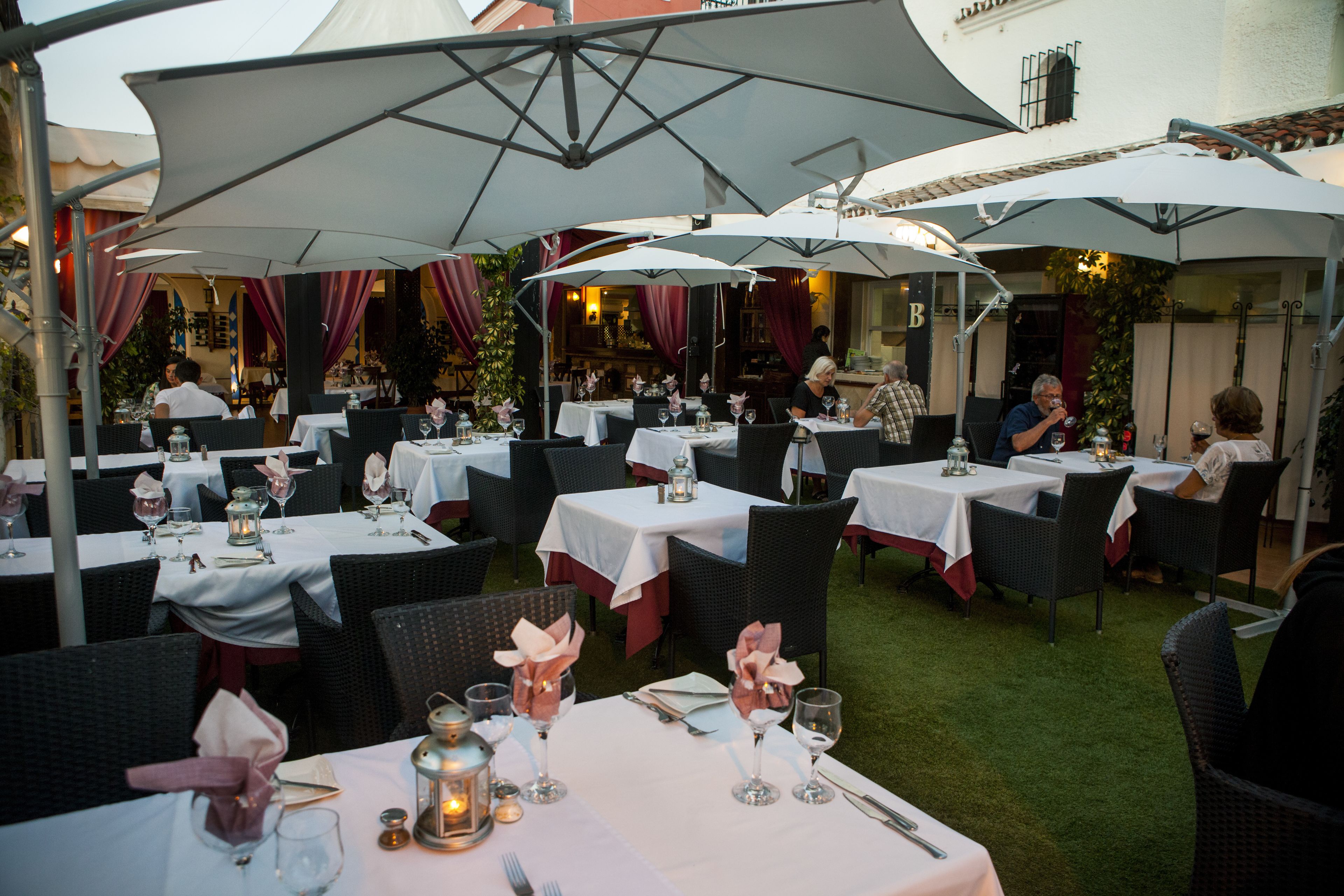 An outdoor dining area with tables set for two, covered by large umbrellas, and surrounded by chairs.