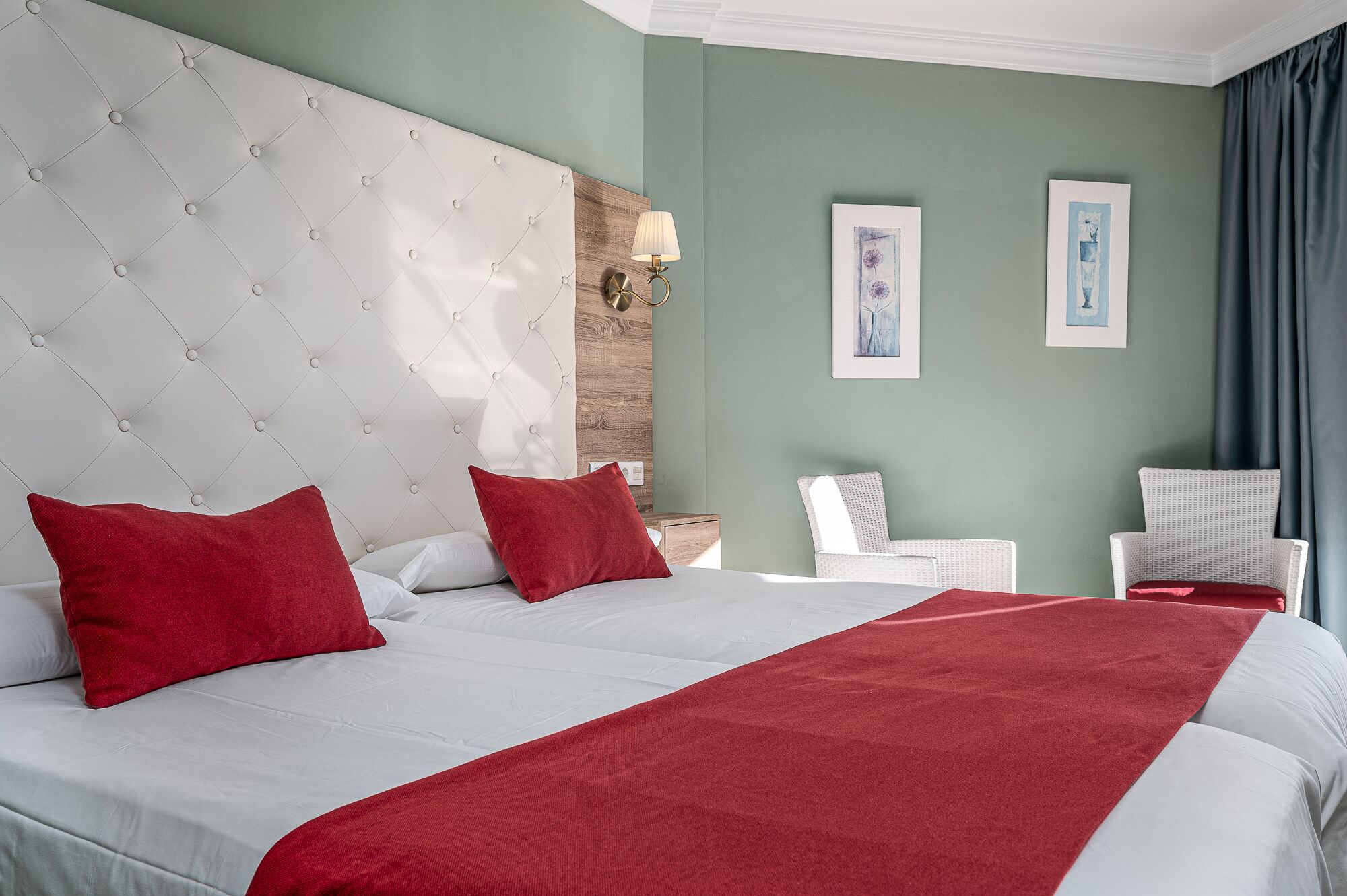 A hotel room with a large bed, two red pillows, a white headboard, a wooden accent wall, a nightstand with a lamp, two framed pictures on the wall, and a window with curtains.