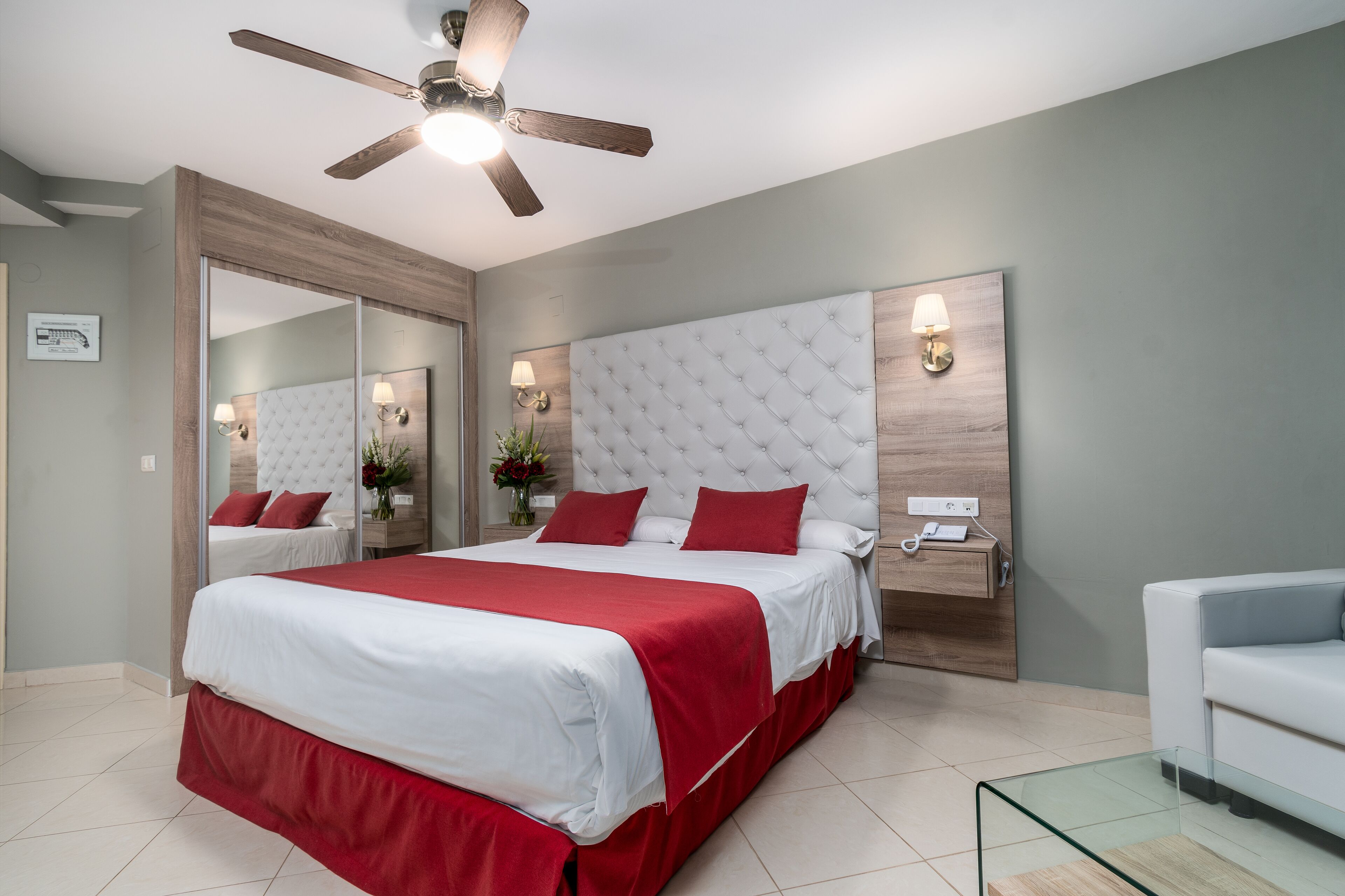 A modern bedroom with a large bed, a ceiling fan, a nightstand with a lamp, and a glass coffee table.