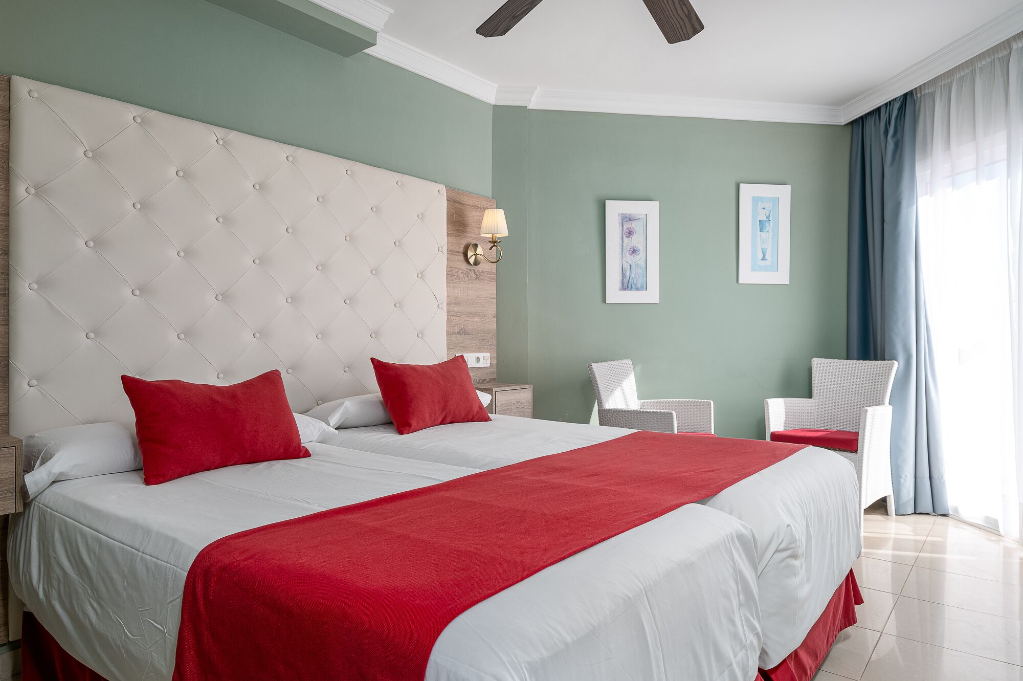 A hotel room with a large bed, two red pillows, a white headboard, and a view of the sea through a window.