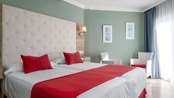 A hotel room with a large bed, two red pillows, a white headboard, and a view of the sea through a window.