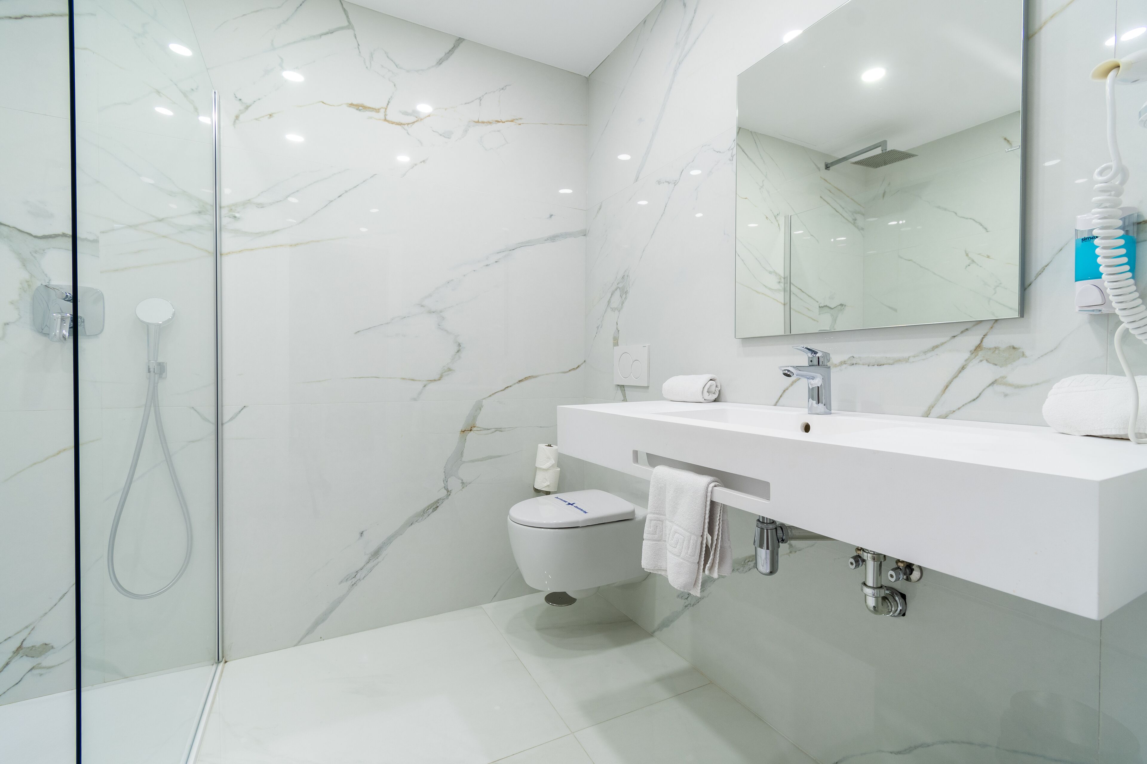 Un baño moderno con pared de mármol, cabina de ducha de vidrio, lavamanos blanco y espejo.