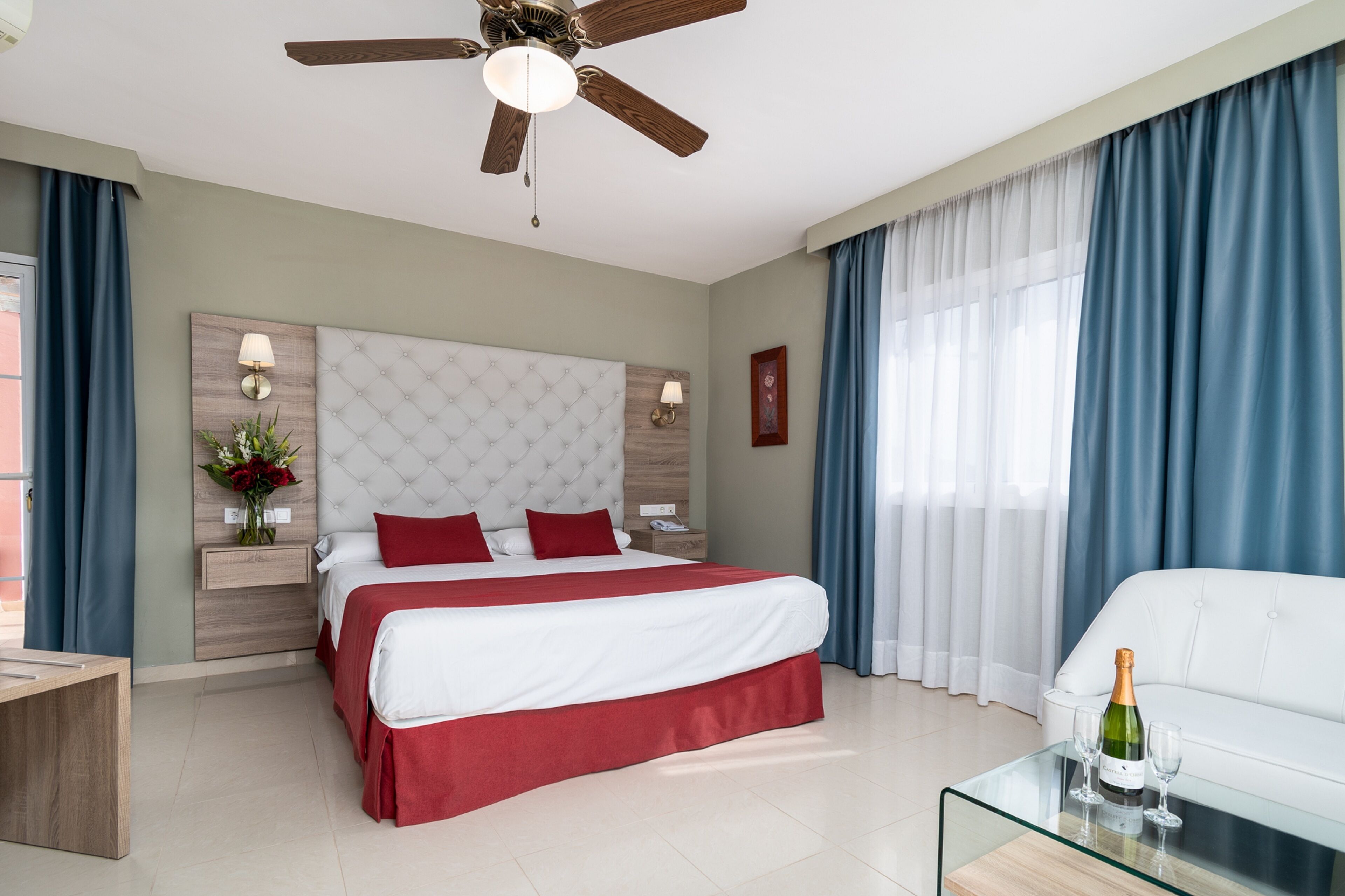 Ein modernes Hotelzimmer mit einem großen Bett, einem Glastisch, einer weißen Couch, einem Deckenventilator und einem großen Fenster mit Vorhängen.