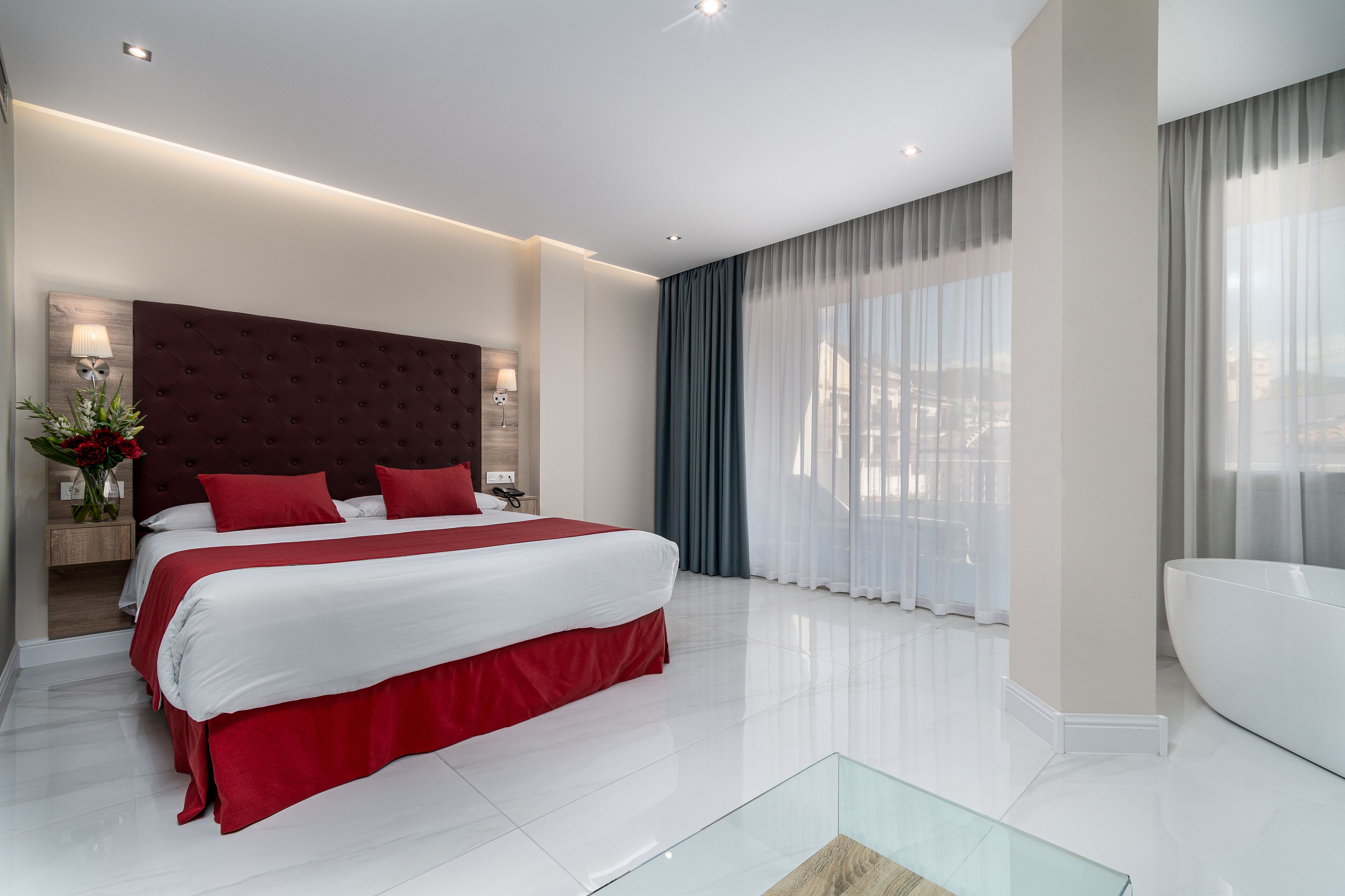 Une chambre d’hôtel moderne avec un grand lit, une table basse en verre, une baignoire blanche et une vue sur la ville.