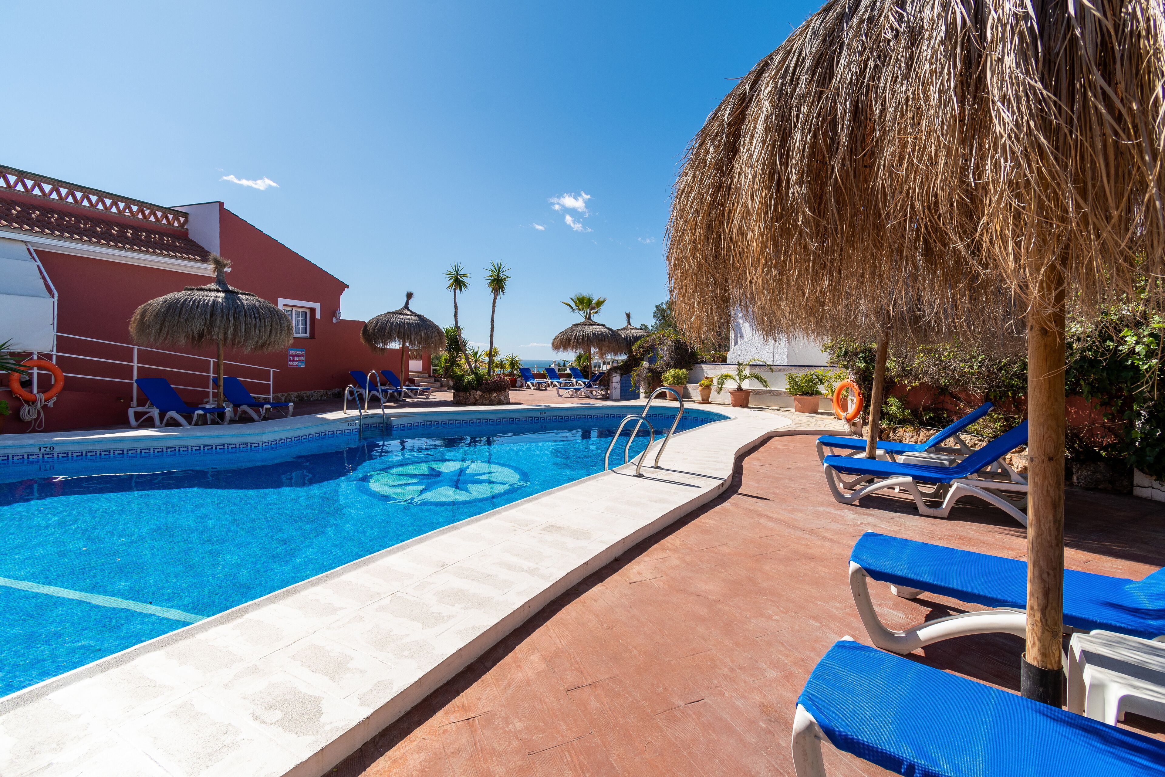 A swimming pool with blue water, surrounded by blue lounge chairs and a red building.