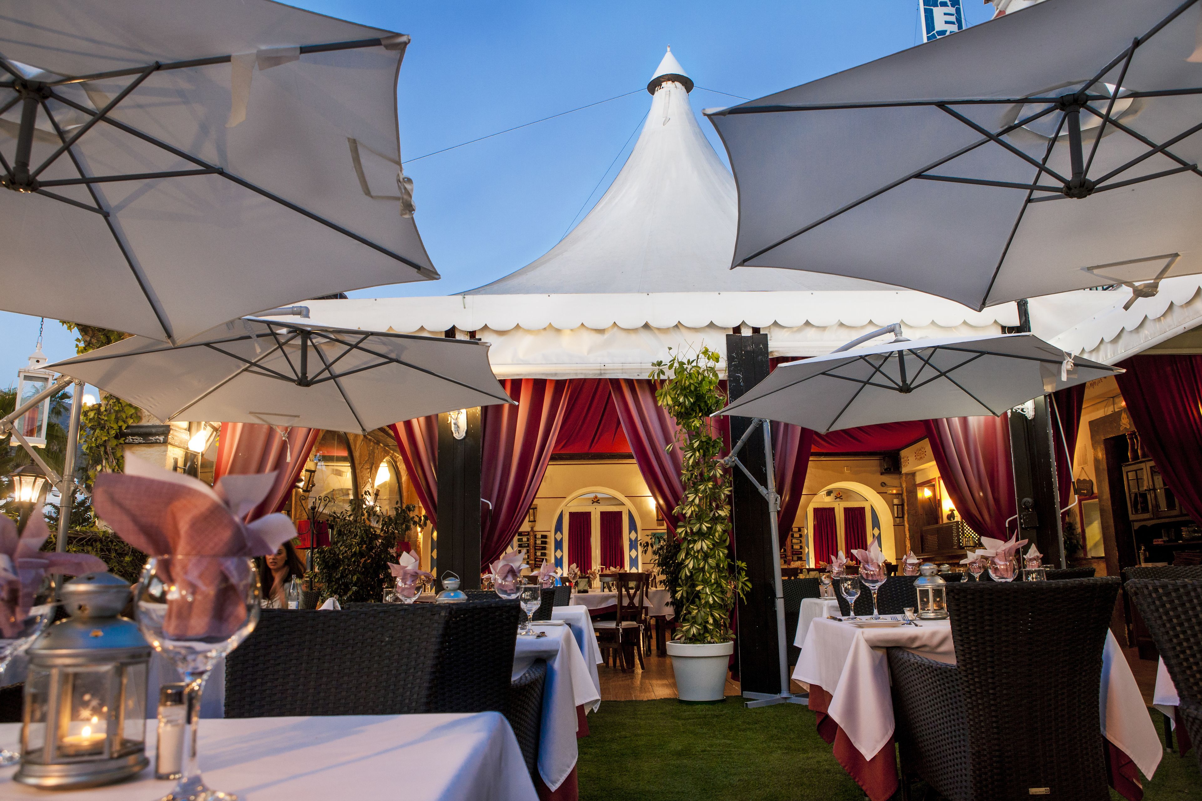 Un área de comedor al aire libre con mesas, sombrillas y cortinas rojas.