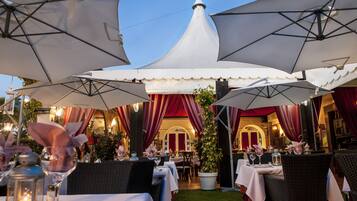 Un área de comedor al aire libre con mesas, sombrillas y cortinas rojas.