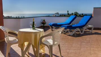 A terrace with a table, two chairs, and two blue lounge chairs facing a view of the sea.