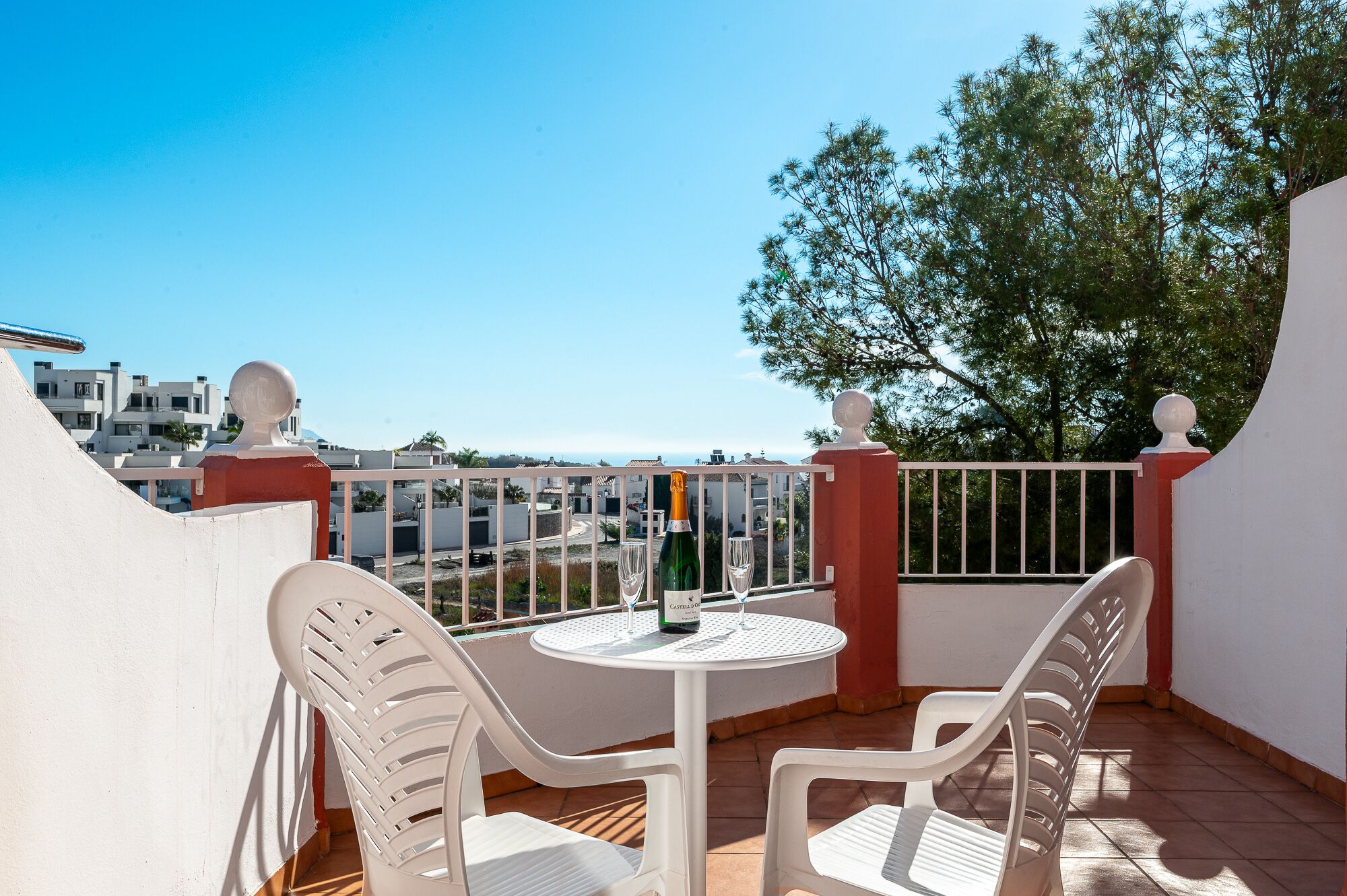 A balcony with a table and chairs, a bottle of champagne, and a view of buildings and trees.