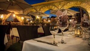 Een buitenterras met tafels gedekt voor een maaltijd, overdekt met grote parasols, en een stenen boog in de achtergrond.