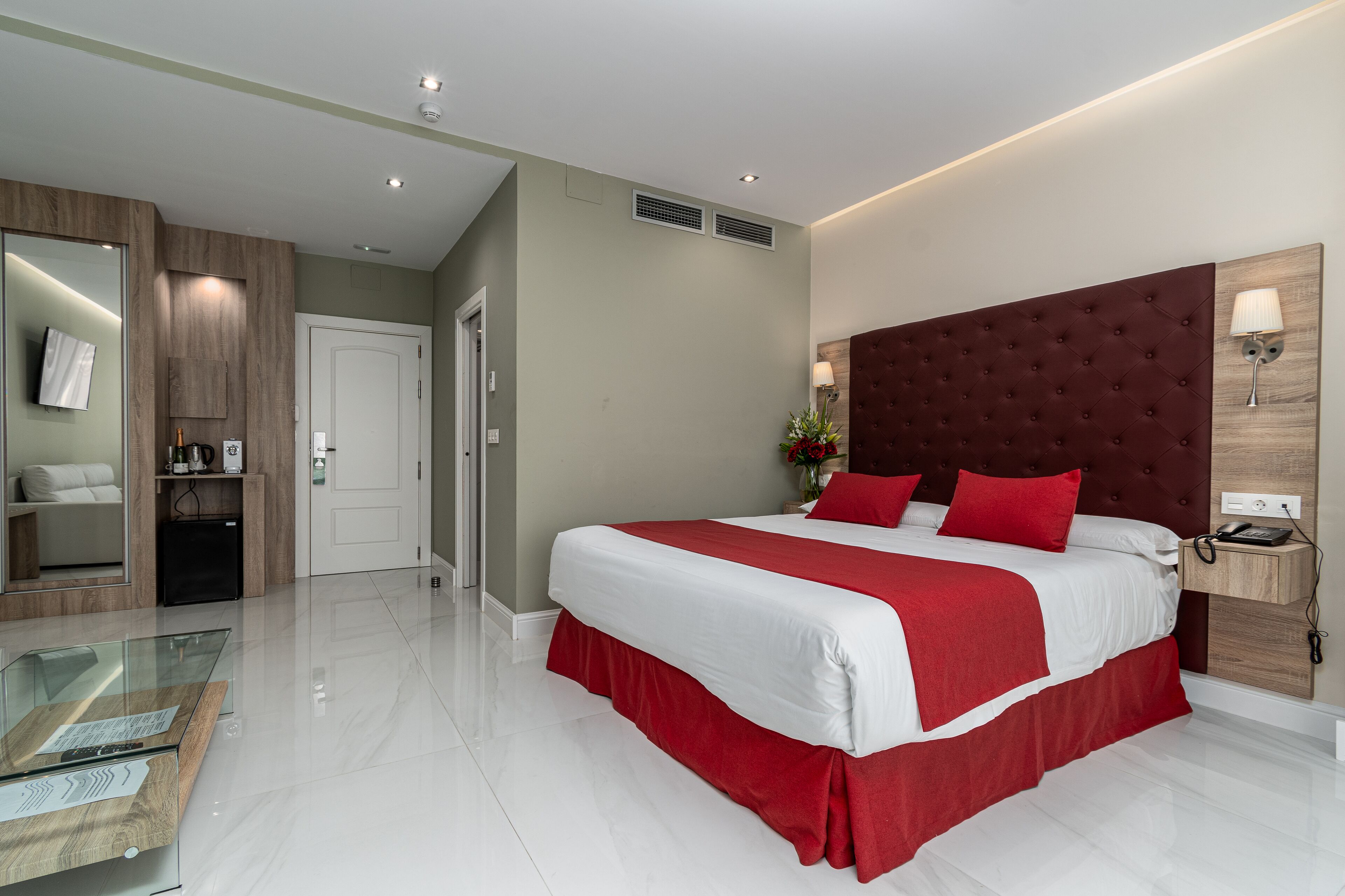 A modern hotel room with a large bed, a glass coffee table, a wooden cabinet, and a mirror.