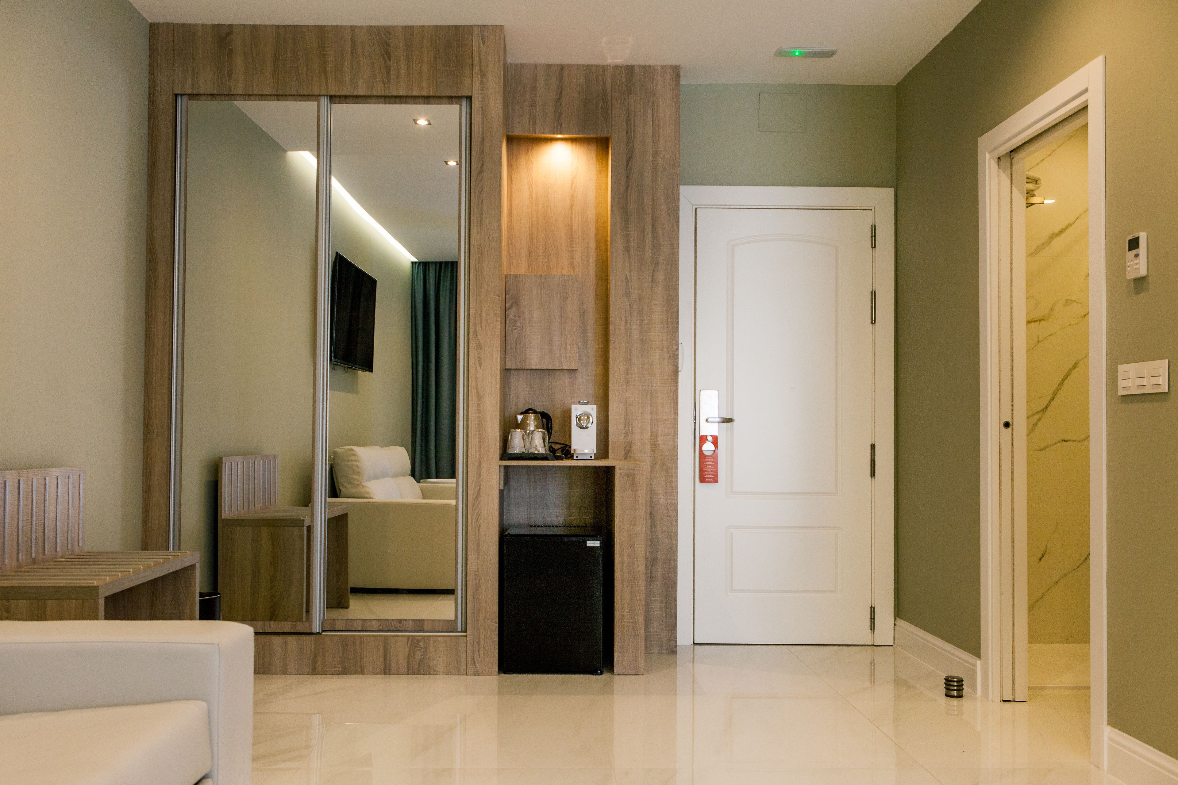 A modern hotel lobby with a mirrored wall, a small refrigerator, a white door, and a marble-patterned wall.